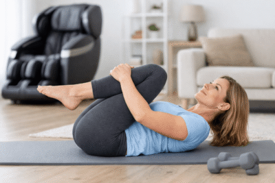 Øvelser til ryggen derhjemme knæ-til-bryst stræk på yogamåtte med massagestol i baggrunden